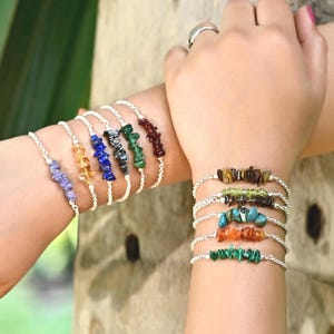 Op de afbeelding: Een verzameling edelsteenarmbanden in verschillende kleuren, waaronder paars, geel, blauw, zwart, groen en rood. De armbanden zijn gemaakt met kleine, onregelmatig gevormde stenen geregen op een zilveren ketting. De armbanden worden op twee polsen getoond.