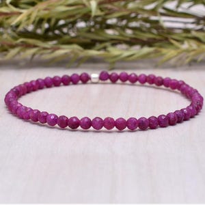 May include: A delicate bracelet featuring small, faceted, magenta-coloured beads strung together. The beads are evenly sized and create a continuous loop. The bracelet is displayed on a light-coloured wooden surface, with greenery in the background.