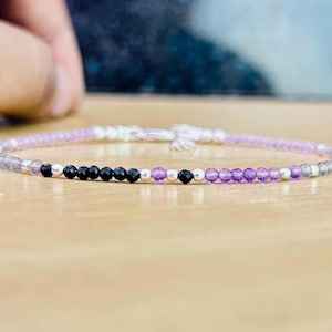 Könnte beinhalten: Ein zartes Armband mit kleinen, runden Perlen in Schwarz, Lila und transparent. Die Perlen sind mit einer silberfarbenen Kette aufgefädelt. Das Armband wird auf einer hellen Holzoberfläche präsentiert.