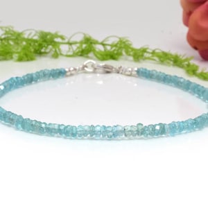 May include: A delicate bracelet featuring small, faceted, light blue gemstones strung together. The bracelet has a silver clasp and is displayed on a white surface with green foliage and a pink flower in the background. The bracelet is a jewelry item.