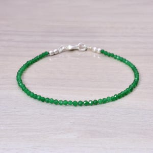 May include: A delicate bracelet featuring small, faceted green beads strung together. The bracelet has a silver clasp and is displayed on a light-colored wooden surface. The beads are a vibrant green color.