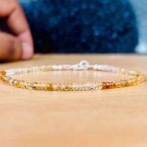May include: A delicate bracelet featuring small, round, amber-colored beads strung together with a silver clasp. The beads transition from light yellow to a deeper orange hue. The bracelet is displayed on a light brown surface.