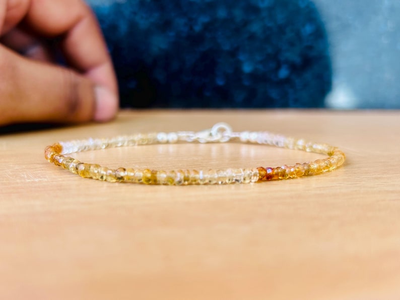 May include: A delicate bracelet featuring small, round, amber-colored beads strung together with a silver clasp. The beads transition from light yellow to a deeper orange hue. The bracelet is displayed on a light brown surface.