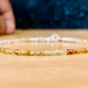 May include: A delicate bracelet composed of small, faceted, amber-colored gemstones. The bracelet features a silver clasp and a gradient of colors, transitioning from light yellow to a deeper orange hue. The gemstones are strung together, creating a subtle, elegant accessory.