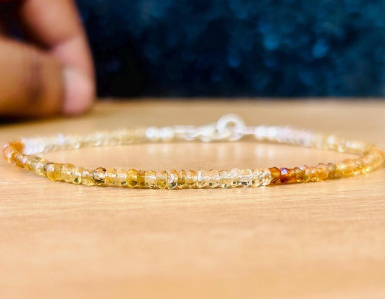 May include: A delicate bracelet composed of small, faceted, amber-colored gemstones. The bracelet features a silver clasp and a gradient of colors, transitioning from light yellow to a deeper orange hue. The gemstones are strung together, creating a subtle, elegant accessory.
