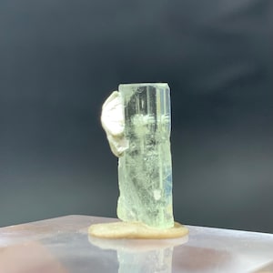 Può includere: Cristallo di tormalina verde pallido con un minerale bianco attaccato. Il cristallo è eretto, di forma rettangolare e con striature verticali. La base è di colore beige chiaro e lo sfondo è scuro.