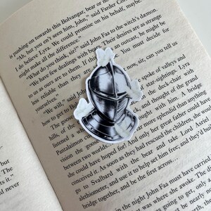 Op de afbeelding: Een sticker met een zwart-witte illustratie van een middeleeuwse ridderhelm omringd door kleine witte motten, geplaatst op een open boekpagina met gedrukte tekst die zinsneden bevat zoals Bolvangar, John Faa en Lyra.