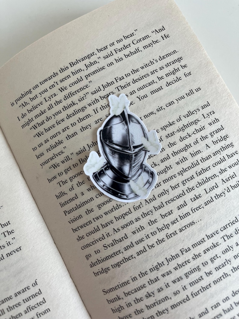 Op de afbeelding: Een sticker met een zwart-witte illustratie van een middeleeuwse ridderhelm omringd door kleine witte motten, geplaatst op een open boekpagina met gedrukte tekst die zinsneden bevat zoals Bolvangar, John Faa en Lyra.