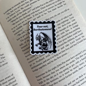 Puede incluir: Una pegatina rectangular con borde negro y bordes festoneados, que presenta una ilustraci&oacute;n en blanco y negro de una g&aacute;rgola posada en una repisa. El texto "Fear not, Bartholomew!" est&aacute; impreso en tinta negra sobre la ilustraci&oacute;n.