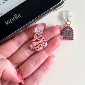 May include: Two small decorative charms with clear plastic clips. One charm is a pink mug shape with the text Witch's Brew and star accents. The other charm is a pink and black arched sign shape with the text Cozy Vibes Only and star accents.