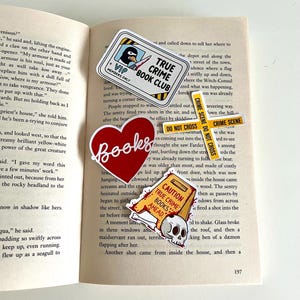 May include: A selection of stickers on an open book. Stickers feature a red heart with "Books", a "True Crime Book Club" badge, a "Crime Scene Do Not Cross" sign, and a caution sign with a skull.
