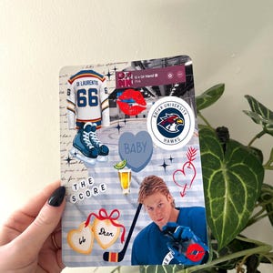 May include: A decorative card featuring hockey-themed graphics. Includes a hockey jersey with the number 66, ice skates, a hockey stick, and a photo of a hockey player. The card also has the text "THE SCORE" and "BABY".