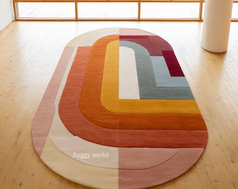 May include: An oval-shaped area rug featuring a geometric pattern of concentric rounded bands in shades of pink, orange, yellow, white, teal, and deep red. The text Ruggy world is printed in white on the lower left section of the rug surface.