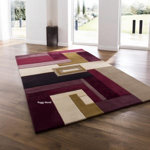 Puede incluir: Una alfombra rectangular con un diseño geométrico en tonos burdeos, beige, dorado y negro. La alfombra presenta un patrón moderno y abstracto con formas rectangulares y cuadradas. La alfombra está colocada sobre un suelo de madera en una habitación bien iluminada.