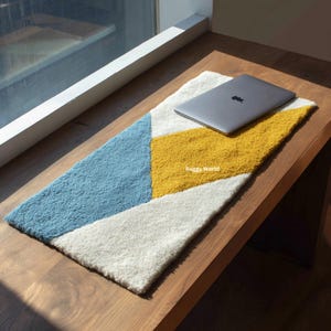 Puede incluir: Una alfombra rectangular con diseño geométrico en azul, amarillo y blanco. Un portátil gris descansa sobre ella, sobre una superficie de madera cerca de una ventana. La alfombra tiene un aspecto texturizado y afelpado. Mide unos 120 cm de largo.