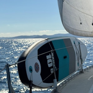 Könnte beinhalten: Ein aufblasbares Stand-Up-Paddleboard mit einem schwarz-weiß-türkisen Design, das auf einem Boot befestigt ist. Das Board trägt den Text "10'6" und ein rundes Logo. Die Szene spielt an einem sonnigen Tag mit dem Meer im Hintergrund.