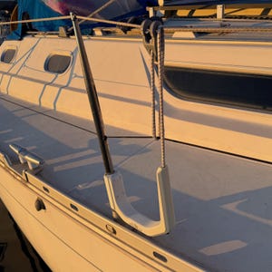 Pode incluir: Um suporte em forma de U para motor de popa montado no corrim&atilde;o de um veleiro. O suporte possui um poste de metal vertical e um cord&atilde;o el&aacute;stico grosso preso a um bloco de polia de metal fixado ao cabo de seguran&ccedil;a horizontal acima.