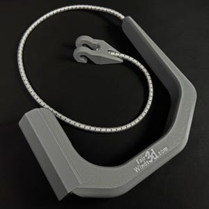 Pode incluir: Uma al&ccedil;a cinza em forma de U com acabamento texturizado e o texto FairWinds3D.com impresso na lateral. Inclui um cord&atilde;o el&aacute;stico flex&iacute;vel prateado e branco preso &agrave; al&ccedil;a, com um pequeno componente de gancho de pl&aacute;stico cinza em forma de V.
