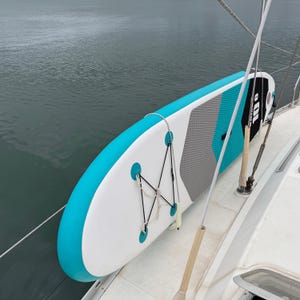 Pode incluir: Uma prancha de stand-up paddle infl&aacute;vel com um esquema de cores branco, turquesa e cinza e um deck pad texturizado. A prancha inclui um sistema de cordas el&aacute;sticas pretas para armazenamento e exibe o texto 10'6 na superf&iacute;cie.