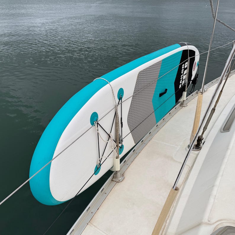 Pode incluir: Uma prancha de stand-up paddle infl&aacute;vel com um padr&atilde;o geom&eacute;trico em branco, azul-petr&oacute;leo e preto, apresentando o texto SAILOR no deck. A prancha est&aacute; presa com cordas brancas ao corrim&atilde;o de metal do conv&eacute;s de um barco sobre &aacute;gua escura.