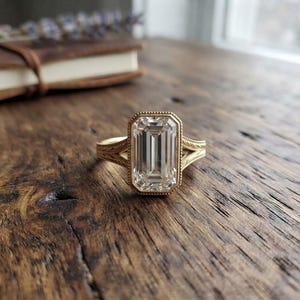 Puede incluir: Un anillo de oro con una gran piedra preciosa transparente de corte esmeralda rectangular engastada en un bisel con cuentas. La banda muestra un patrón detallado grabado en forma de hoja en los hombros, desde el engaste central.
