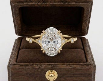 May include: A yellow gold ring featuring a large oval-cut center stone held by prongs, with smaller round accent stones set along the band. The ring is displayed inside a dark brown wooden box with a velvet interior and a gold-toned latch.