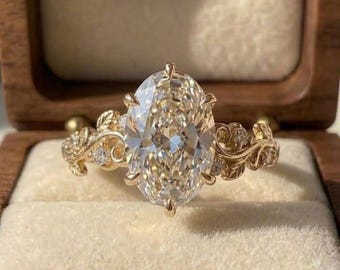 Könnte beinhalten: Ein goldener Ring mit einem großen oval geschliffenen Mittelstein, der von Krappen gehalten wird. Die Ringschiene ist mit filigranen Metallarbeiten in Form von Blättern und Ranken verziert, besetzt mit kleinen runden Steinen.