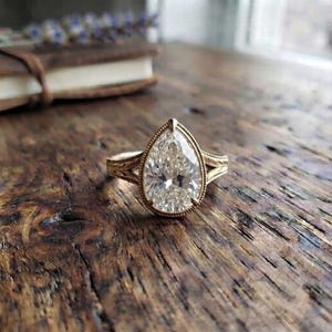 May include: A gold ring featuring a large, pear-shaped, faceted clear gemstone set in a bezel setting. The band of the ring displays a detailed, engraved leaf-like pattern that extends from the central stone along the sides of the metal band.