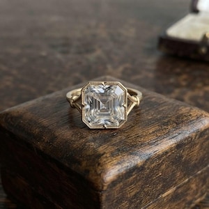 Puede incluir: Un anillo de oro con una piedra preciosa transparente de talla cuadrada y facetada, engastada en un marco con borde de cuentas. La banda se divide en dos brazos finos y angulados que se conectan al engaste central, creando un diseño geométrico.