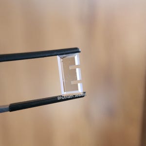 以下が含まれることがあります： 黒いピンセットで保持された、透明な長方形の宝石。宝石は「E」の形をしており、透明です。「Bhutnath Jewels」の文字が宝石に印刷されています。