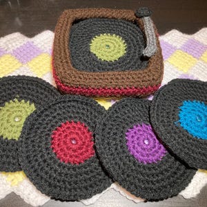 May include: A crocheted coaster set depicting a record player and vinyl records. The record player is brown, black, and grey, with a green centre. The vinyl records are black with various coloured centres: green, red, purple, and blue. The set is on a crocheted placemat.