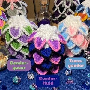 Op de afbeelding: Gehaakte bollen met schubachtige patronen in verschillende kleuren en witte toppen. Eén bol heeft paarse, blauwe en roze schubben. De afbeelding bevat de tekst "Gender-queer", "Gender-fluid" en "Transgender".