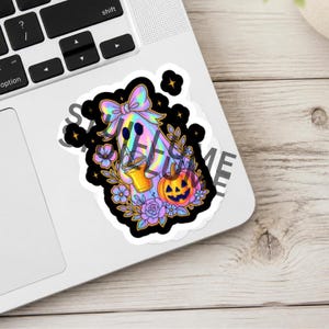 May include: A holographic sticker featuring a ghost with a large bow, holding an orange drink, surrounded by purple flowers and a jack-o'-lantern. The design includes small yellow stars against a black background, applied to a laptop.