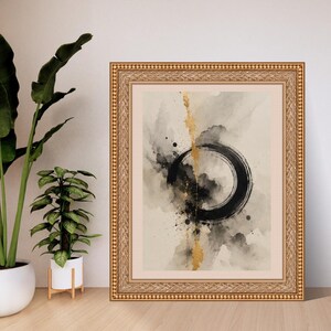 Op de afbeelding: Een ingelijste wandkunstprint met een zwarte cirkelvormige penseelstreek in het midden op een beige achtergrond, met abstracte grijze aquarelvegen en een verticale streep van bladgoudtextuur door het midden van de compositie.