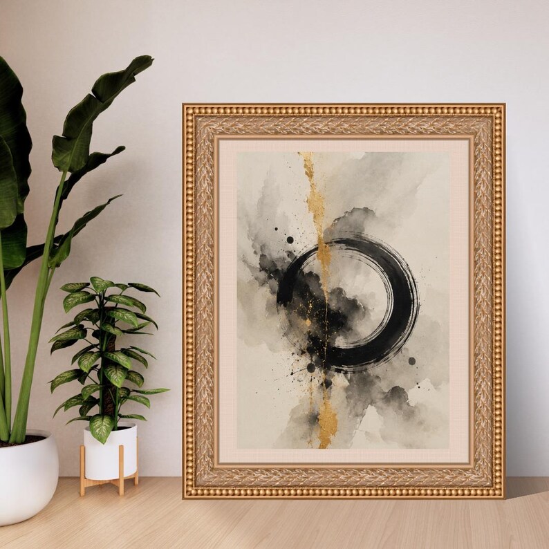 Op de afbeelding: Een ingelijste wandkunstprint met een zwarte cirkelvormige penseelstreek in het midden op een beige achtergrond, met abstracte grijze aquarelvegen en een verticale streep van bladgoudtextuur door het midden van de compositie.