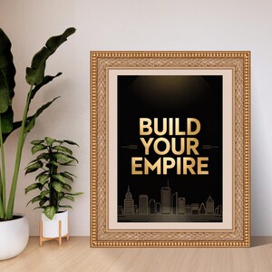 May include: A framed wall art piece featuring a black background with the gold text BUILD YOUR EMPIRE centered above a gold line-art city skyline. The artwork is enclosed in an ornate, textured gold frame with a light-colored matting border.