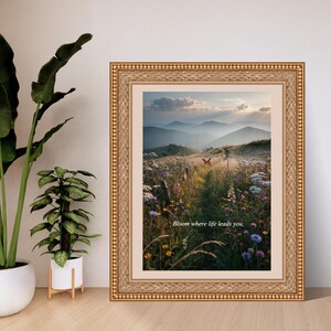 May include: A framed wall art print featuring a landscape of a wildflower meadow at sunset with distant mountains and a butterfly. The text "Bloom where life leads you." is printed in the center. The frame is ornate, metallic gold with a textured border.