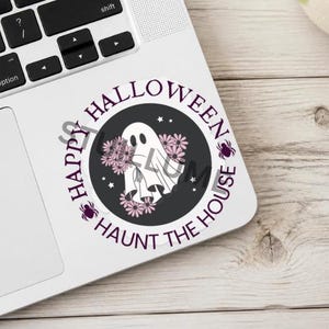 Puede incluir: Una pegatina circular con un fantasma blanco rodeado de flores rosas sobre un fondo gris oscuro. El texto "HAPPY HALLOWEEN" y "HAUNT THE HOUSE" est&aacute; impreso en morado alrededor del borde, con peque&ntilde;as estrellas y dos ara&ntilde;as moradas.