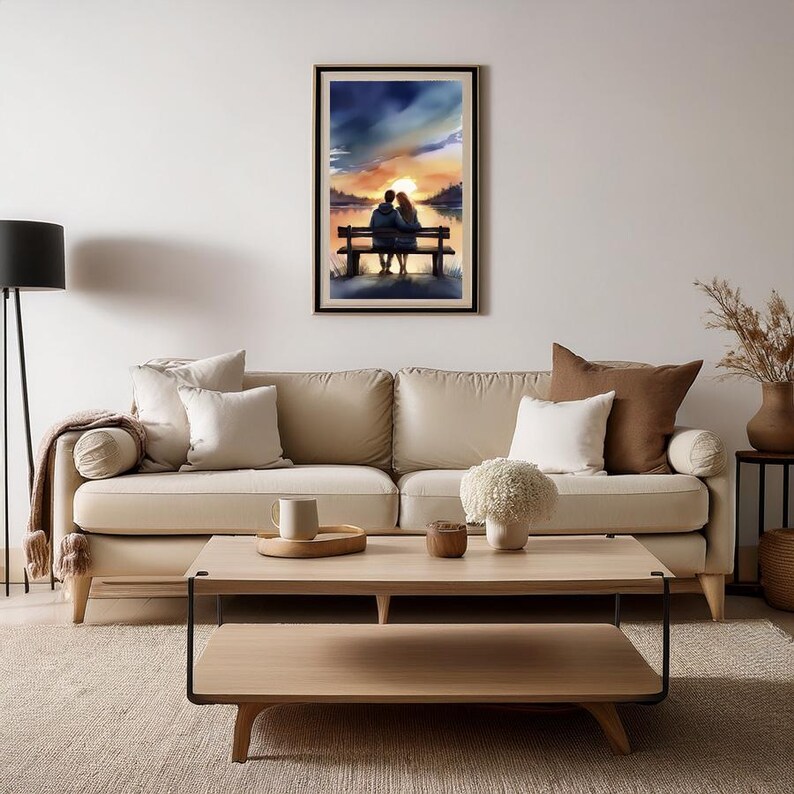 May include: A framed watercolour painting shows a couple on a bench, silhouetted against a sunset over a lake. The artwork uses blues, oranges, and yellows, creating a romantic and serene atmosphere. The painting is displayed above a beige sofa.