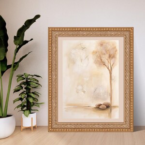 K&ouml;nnte beinhalten: Ein gerahmtes Wandbild mit einem abstrakten Aquarellgem&auml;lde eines einzelnen Baumes und drei glatten Steinen in Beige-, Creme- und Braunt&ouml;nen. Das Kunstwerk befindet sich in einem verzierten, strukturierten Goldrahmen mit einem breiten, neutralen Passepartout.