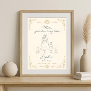 May include: Framed print with a beige background featuring a mother and child illustration. The text reads "Mom, your love is my home" and "Sophia, Love, Emma, Mother's Day 2026". The frame is a light brown colour.