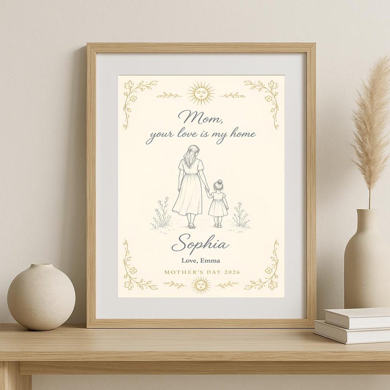 May include: Framed print with a beige background featuring a mother and child illustration. The text reads "Mom, your love is my home" and "Sophia, Love, Emma, Mother's Day 2026". The frame is a light brown colour.