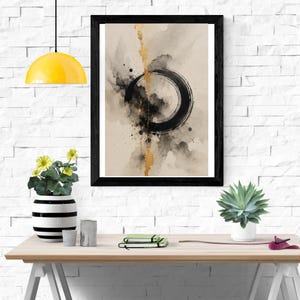 Op de afbeelding: Een ingelijste wandkunstprint met een abstract aquarelontwerp met een opvallende zwarte cirkelvormige penseelstreek, een verticaal bladgoudaccent en donkere inktspatten op een neutrale beige achtergrond, geplaatst in een eenvoudige zwarte lijst.