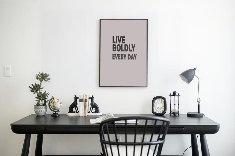 May include: A modern home office setup with a black desk, chair, and accessories. A framed print with the text "LIVE BOLDLY EVERY DAY" hangs above the desk. Decorative items include a potted plant, globe, bookends, and a desk lamp.