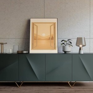 May include: A dark teal modern sideboard with gold legs, holding a framed landscape art print. A small potted plant and a table lamp are on the sideboard. The art print depicts a sunset scene with an open window.
