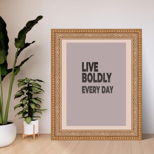 May include: A framed print with the text "LIVE BOLDLY EVERY DAY" in black letters. The frame is gold with a detailed pattern. The background of the print is a muted purple color. Two potted plants are in the background.