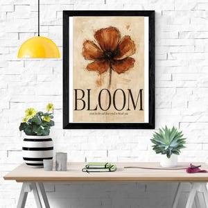 May include: A framed wall art print featuring a large, painterly rust-coloured poppy flower on a beige background. Below the flower, the word BLOOM is printed in large black capital letters, followed by the text even in the soil that tried to break you.