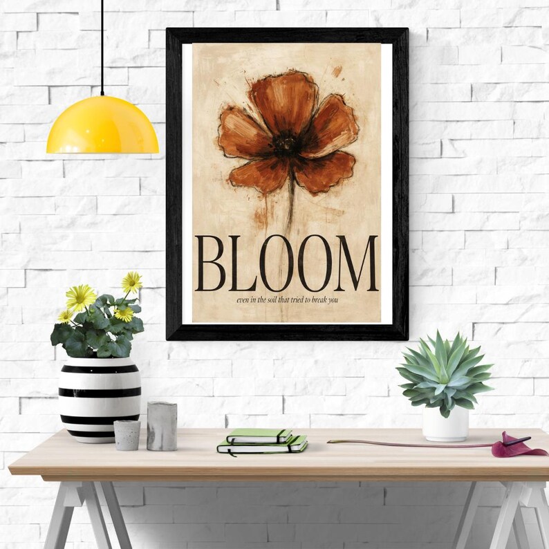 May include: A framed wall art print featuring a large, painterly rust-coloured poppy flower on a beige background. Below the flower, the word BLOOM is printed in large black capital letters, followed by the text even in the soil that tried to break you.