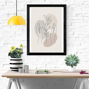 Pu&ograve; includere: Una stampa artistica da parete incorniciata con un disegno a linee nere di due mani che avvolgono un gambo di pianta centrale con foglie. Il design &egrave; su uno sfondo beige chiaro con morbide forme astratte grigie e marroni dietro le mani.