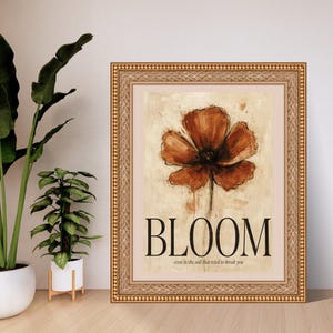 May include: A framed wall art print featuring a painterly rust-coloured flower on a beige background. The text "BLOOM" is printed in large black capital letters below the flower, with the smaller text "even in the soil that tried to break you" underneath.